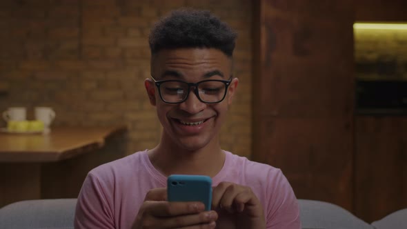 20s African American Man Wearing Eyeglasses Using Cell Phone and Surfing Internet Sitting on Sofa at alt