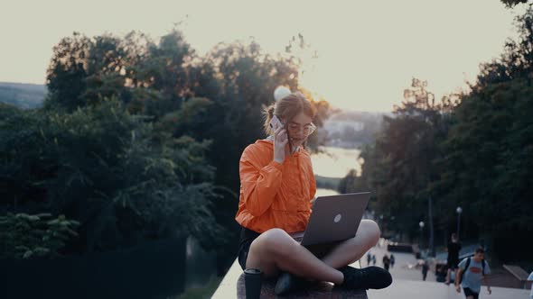 The Serious Young Woman Who Works on the Laptop Answers the Phone Starts to Smile Widely Hears alt