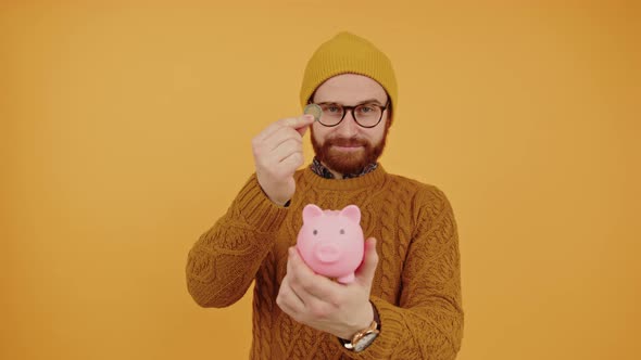 Smiling Millennial Caucasian Hipster Man Putting Money to His Piggy Bank Yellow Studio Background alt