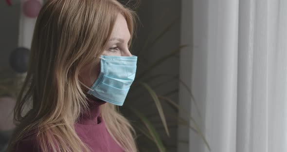 Side View Close-up of Sad Caucasian Woman in Face Mask Looking Out the Window on Covid-19 Lockdown alt