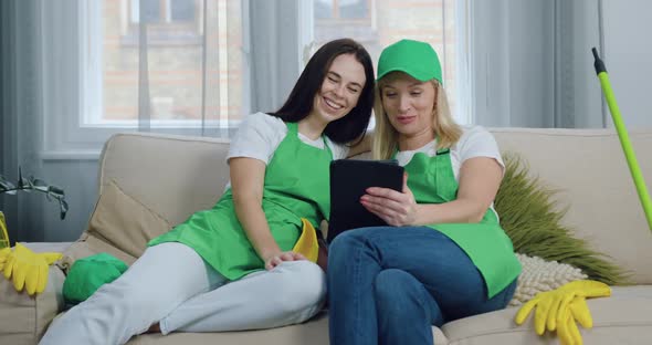 Two Women-Cleaners Resting on Couch in Contemporary House and Watching Funny Videos on Tablet PC alt