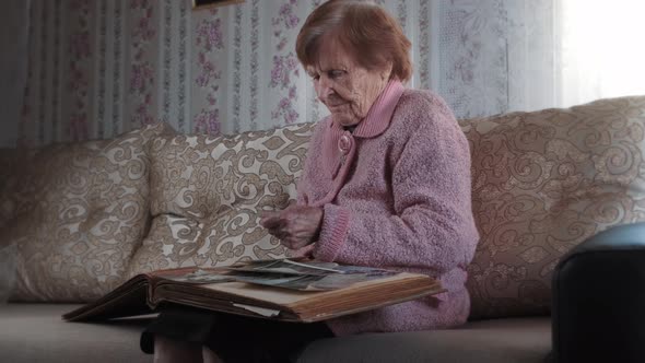 Old grandmother holds and looks at photographs with nostalgia, sitting on the sofa alt