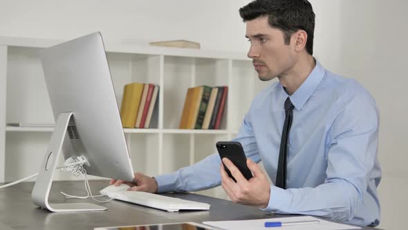 Businessman Using Smartphone While Working on Computer alt