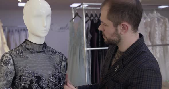 Tailor Corrects Sewn Dress on a Mannequin and Looks at Camera with Crossed Hands alt
