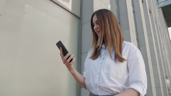 Young Woman in White Shirt Surfing the Internet on the Background of the Business Center Sunny Day alt