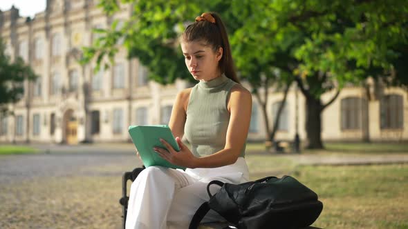 Young Thoughtful Woman Surfing Internet on Tablet Sitting on Bench on Sunny University Yard Looking alt
