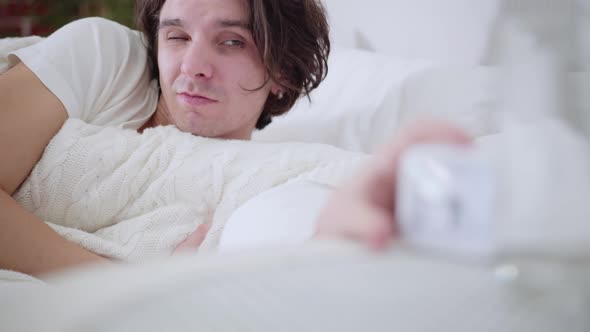 Closeup Portrait of Sleepy Young Man Looking at Alarm Clock and Waking Up with Shocked Facial alt
