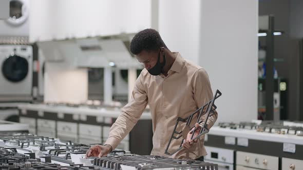 Black Man with Face Mask is Choosing Stove for New Kitchen in Apartment Shopping in Home Appliances alt