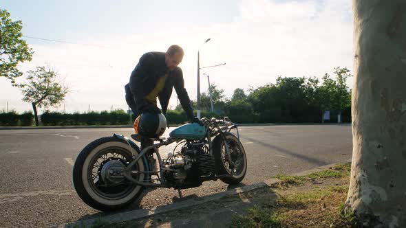 Young Stylish Motorcyclist Goes to His Custom Bobber Motorcycle on Street Road at Sunset Slow Motion alt