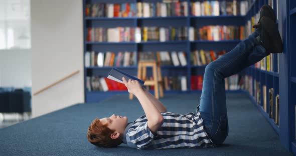 Redhead Preteen Boy Lying on Floor and Reading Book in Library alt