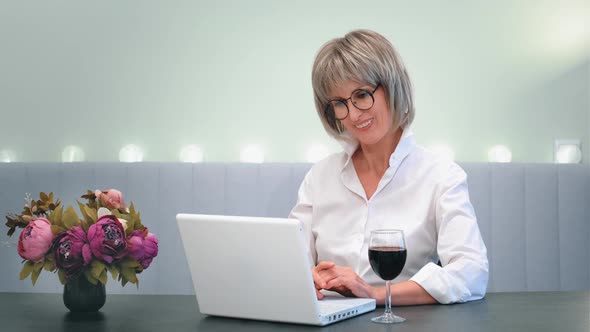 A Happy Beautiful Elderly Lady Drinking Wine Calls a Friend a Relative From Home on a Laptop Video