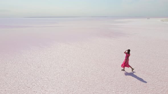 Cinematic Aerial Drone Following Attractive Young Woman in Pink Dress Walking on Colorful Pink Lake alt