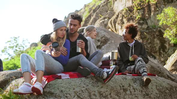 Beautiful Pair Sitting on Plaid on Rocks with Thermocups While Their Interracial Friends Chatting alt