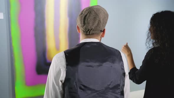 Two Visitors of Modern Art Museum Are Sitting on Bench in Front Abstract Picture alt