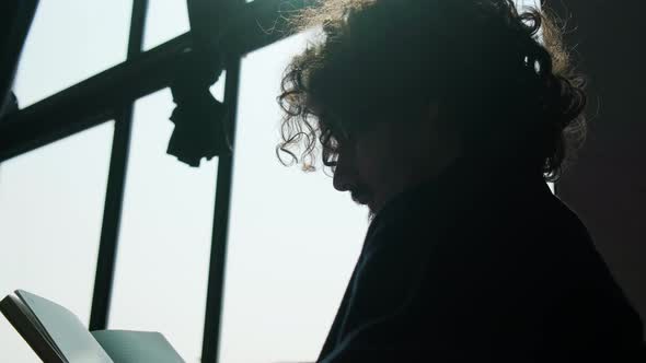 Silhouette of a Man Sitting Near Window and Reading a Book at the Sunset alt