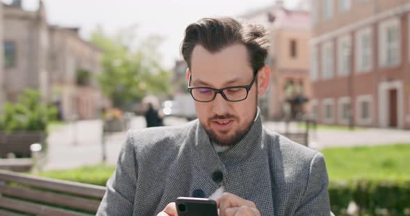 Young Man Wearing Glasses with Mustaches and a Beard is Sitting on a Bench in the Square Reading alt