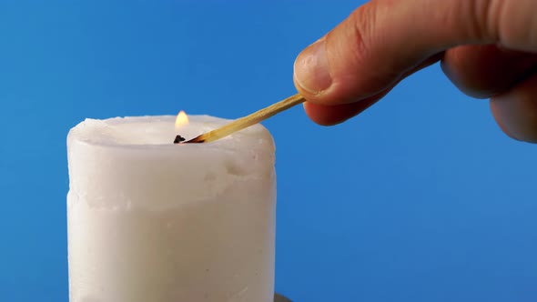 Hand Lights White Wax Candle Using a Match on a Blue Background alt