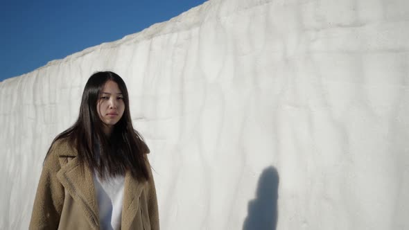 Thoughtful Beautiful Asian Woman Walks Along a Snowy Wall Squinting From the Sun
