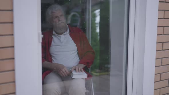 Depressed Thoughtful Senior Handicapped Man Closing Book Looking Out Window on Glass Door alt