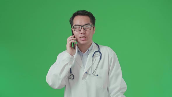 Asian Doctor With Stethoscope Talking On Mobile Phone While Walking On Green Screen alt