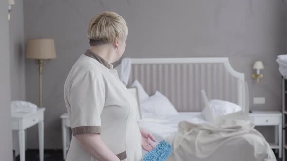 Exhausted Professional Maid Looking Back at Bed in Hotel Room and Turning To Camera. Middle Shot alt