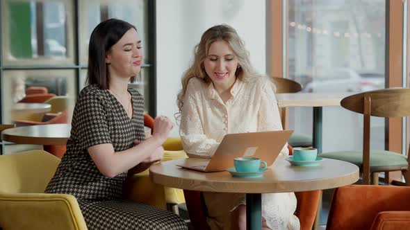 Cheerful Carefree Young Women Surfing Social Media on Laptop and Gossiping Sitting in Cafe Indoors alt