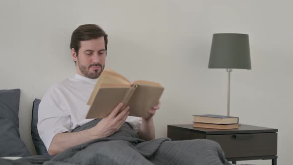 Young Man Reading Book While Sitting in Bed alt