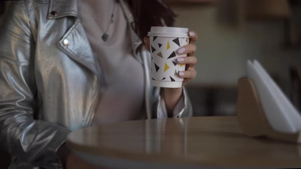 A Woman in a Silver Jacket Sits in a Coffee Shop with a White Cup of Coffee