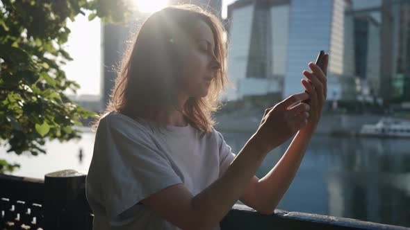 a Woman Leans on the Fence and Attentively Looks at the Phone Screen on the Embankment Against the alt