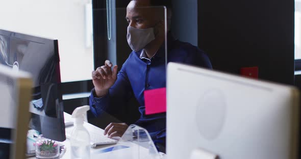 African american man wearing face mask using computer while sitting on his desk at modern office alt