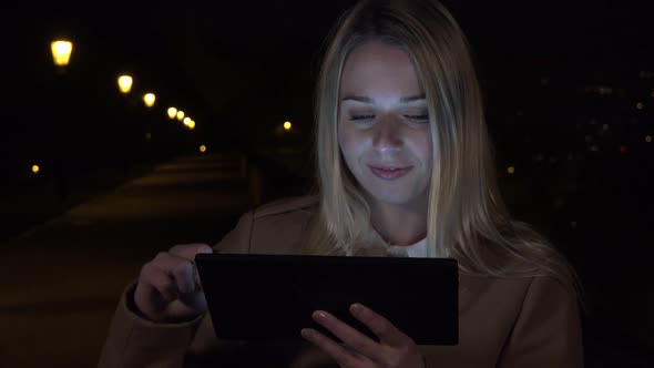A Young Beautiful Woman Works on a Tablet with a Smile in an Urban Area at Night alt