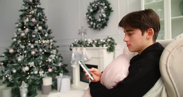 Young Man Browsing Phone on Sofa, Christmas Tree in Background alt