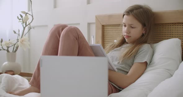 Tired and Bored Pupil Girl Is Lying on Bed with Exercise Book and Listening Lesson on Laptop, e alt