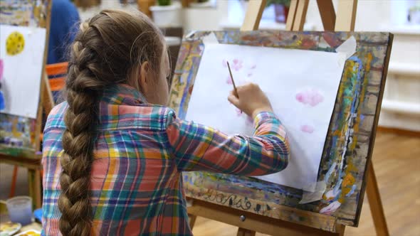 Young Beautiful Female Artist is in an Art Studio, Sitting Behind an Easel and Painting on Canvas. alt
