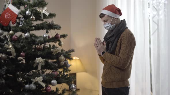 Side View of Hopeful Young Man in Covid19 Face Mask Praying To Christmas Tree at Home alt