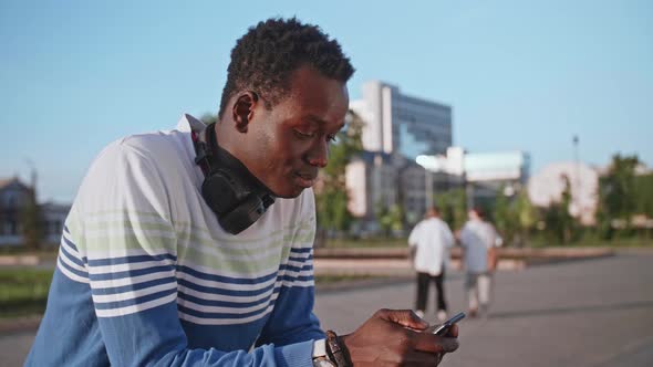 A Young Black Student in Stylish Clothes Sits and Talks on a Smartphone in the City