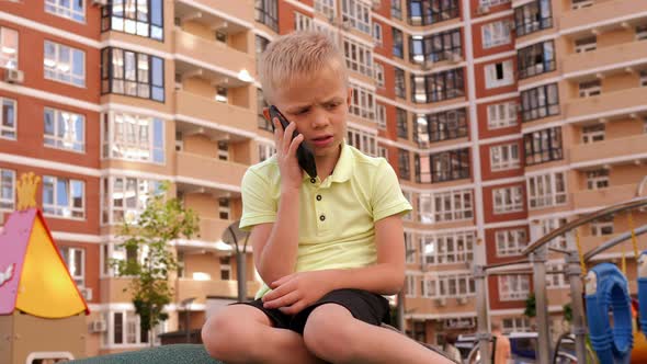 A Little Sad Boy is Talking on the Phone in the Playground alt