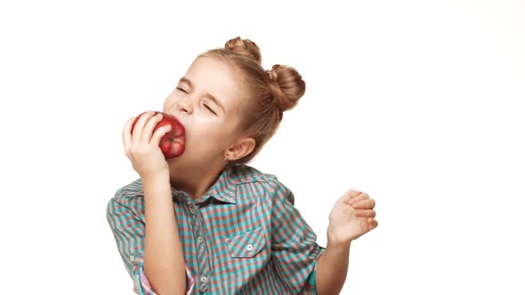 Small Smiling Caucasian Kid Girl with Hair Buns and Plaid Shirt Trying to Bite Red Apple on White