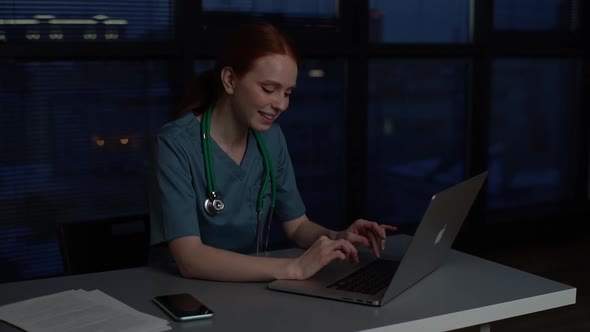 Positive Cheerful Redhead Young Female Doctor in Blue Green Medical Uniform Working Typing on Laptop alt