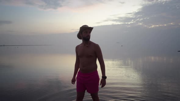 Adult Bearded Man in Sunhat Turning Around Amazed with Picturesque Seaside Sunset and Sunlight alt