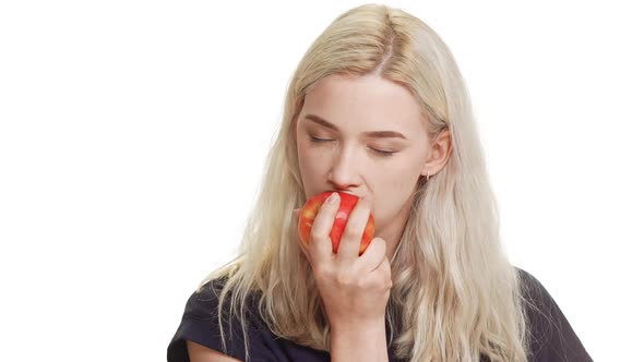Charming Caucasian Blonde Teenage Girl in Dark Blue Tshirt Biting Eating Apple on White Background alt