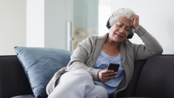 African american senior woman wearing headphones using smartphone sitting on the couch at home alt