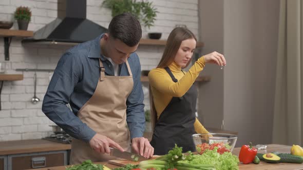 Happy couple enjoying cooking time together at home. Side view of young man cutting vegetables alt