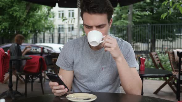Man Drinking Coffee and Using Smartphone While Sitting in Cafe Terrace alt