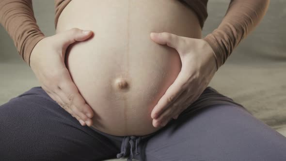 Young Pregnant Woman Stroking Big Belly on Couch Making Heart Shape with Hands alt