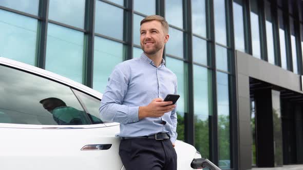 Charging an Electric Car and Man Standing Near the Vehicle and Using His Mobile Phone alt