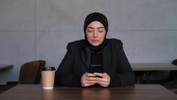 Successful and Serious Young Muslim Woman in Hijab with Smartphone and Coffee Cup Planing Job