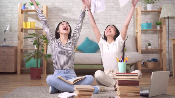 Two Positive Asian Students Are Sitting on the Floor Throwing Papers Up alt