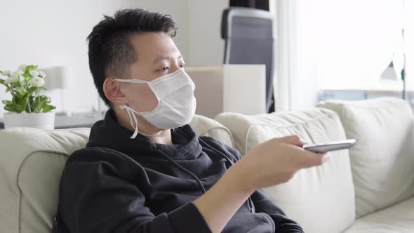 A Young Asian Man in Face Mask Switches Channels with a Remote Control on a TV That's Off the Camera alt
