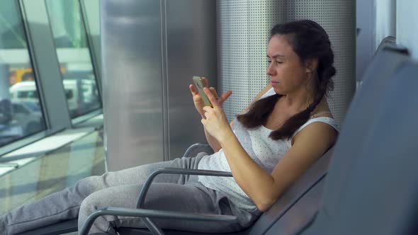 Adult Brunette Woman Is Sitting Alone in Waiting Area in Station, Using Smartphone alt